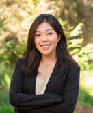 A young asian woman with mid-length dark brown hair wears a cream-colored bouse and navy suit jacket, crosses her arms and smiles at the camera in front of a lush greeen background.